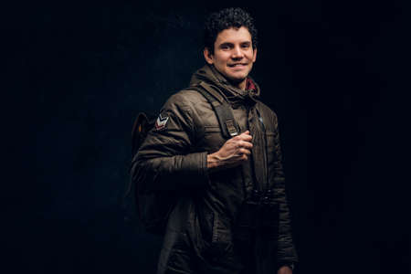 Young handsome traveler with a backpack and binoculars smiling and looking into the camera on black...