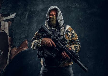 Special forces soldier in military uniform wearing mask and hood holding an assault rifle. Studio photo against a dark textured wallの写真素材