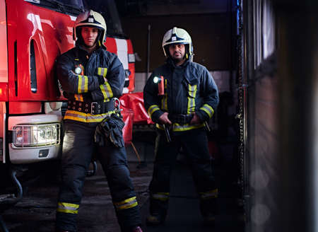 Two firemen wearing protective uniform standing next to a fire engine in a garage of a fire department.の写真素材