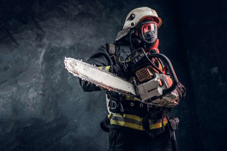 Firefighter in safety helmet and oxygen mask holding a chainsaw. Studio photo against a dark...