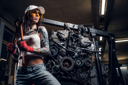 Sexual tattoed girl wearing cap and dirty clothes posing next to a car engine suspended on a hydraulic hoist in the workshopの写真素材