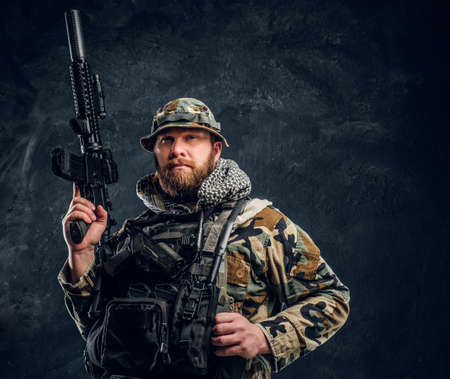 Bearded special forces soldier in the military camouflaged uniform holding an assault rifle. Studio photo against a dark textured wallの写真素材