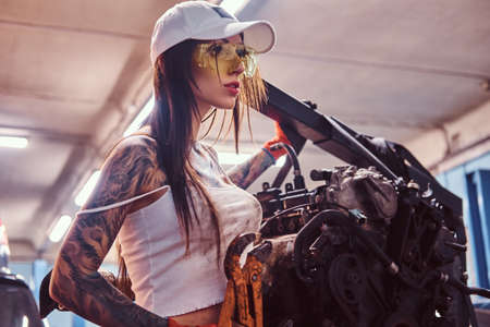 Female car mechanic holding a big hammer and posing next to a car engine suspended on a hydraulic...