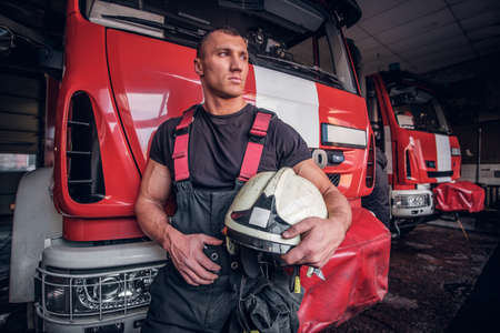 Muscular fireman holding a protective helmet in a garage of a fire department, leaning on a fire...