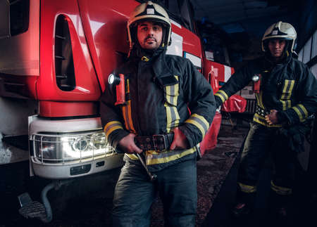Two firemen wearing protective uniform standing next to a fire engine in a garage of a fire...
