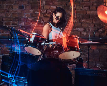 Beautiful girl musician in sunglasses holding the sticks sitting behind the drum set against a brick wall. Perform in a night clubの写真素材