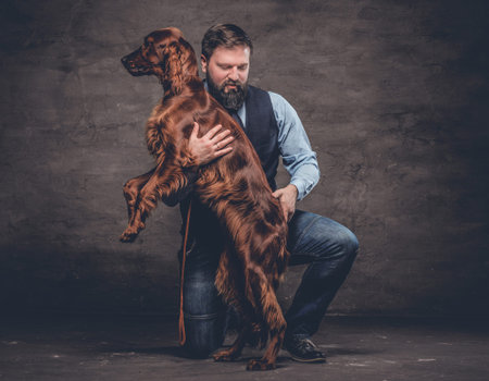 Middle-aged hunter dressed in elegant clothes sits on his knee and holding his purebred brown...
