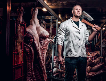 The butcher in a white work shirt holding ax while standing in a refrigerated warehouse in the midst of meat carcassesの写真素材