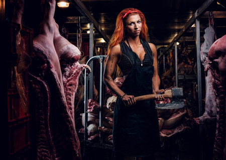 Strong female butcher in workwear posing with ax in a refrigerated warehouse in the midst of meat carcassesの写真素材