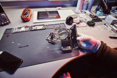 The technician uses a magnifying glass to carefully inspect the internal parts of the smartphone in...