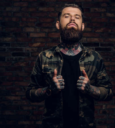 Confident bearded guy with tattoos on his hands and neck in the military shirt. Studio photo against a brick wallの写真素材