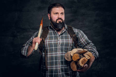 Portrait of a bearded woodcutter with a backpack dressed in a plaid shirt holding firewood and ax. Studio photo against a dark textured wallの写真素材