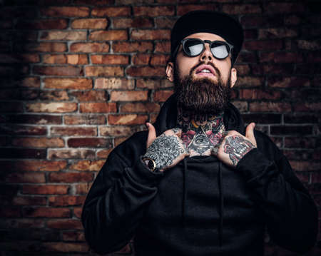 A stylish tattooed guy in a black hoodie and sunglasses. Studio photo against a brick wallの写真素材