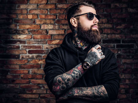 A stylish tattooed guy in a black hoodie and sunglasses. Studio photo against a brick wallの写真素材