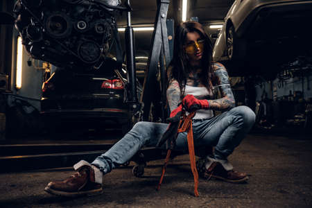 Stylish tattooed girl holding a big wrench while sitting on a hydraulic hoist with a suspended car engine in the workshopの写真素材