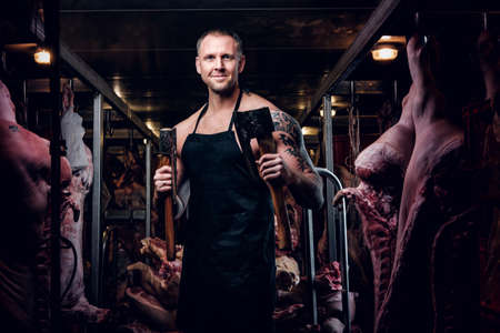 Smiling butcher with a tattoo on his hand wearing apron posing with two axes in a refrigerated warehouse in the midst of meat carcassesの写真素材