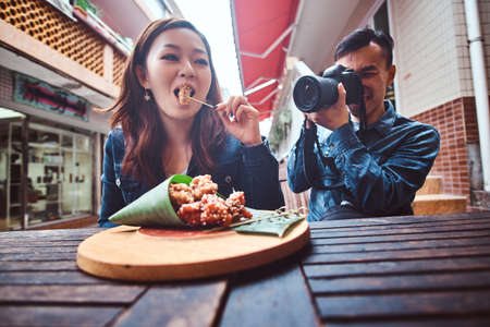 Young asian couple enjoying chinese dish. At front on wooden table is traditional asian food - bamboo leaf wrap. Man is taken a photo of a woman. Woman is trying asian dish at first time. Widescreen shooting.の写真素材