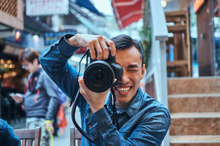 Casual young asian men is making a photo with photo camera outside in public place. He is smiling. Man is wearing denim jacket.の写真素材