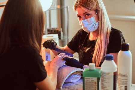 Master manicurist is looking at camera while working with clients nails at the beauty salon. Photo with warm light.の写真素材