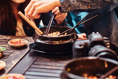 People are enjoying asian food. There is wooden table. On the table are photo camera, traditional asian bowls with food, chopsticks and sauce.の写真素材
