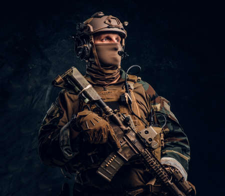 Private security service contractor in camouflage uniform posing with assault rifle. Studio photo against a dark textured wallの写真素材
