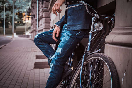 Trendy man in denim is standing on the street. He is leaning on his bicycle. Building and road at the background.の写真素材