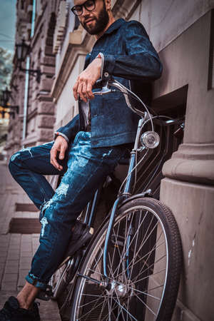 Trendy man in denim and glasses is standing on the street. He is leaning on his bicycle. Man is looking down. Building at the background.の写真素材