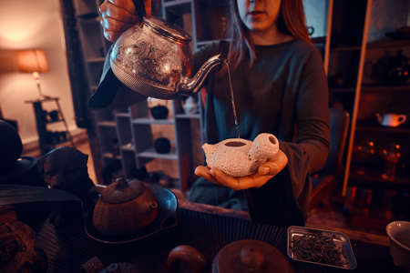 Oriental master wearing a gray dress holding a kettle and pouring natural tea into a teapot in the dark room with a wooden interior. Tradition, health, harmony. Chinese tea ceremonyの写真素材