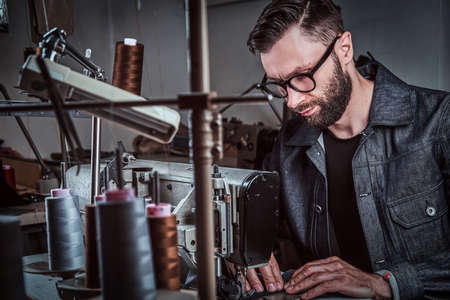 Fashion designer is sewing clothes by vintage sewing machine. He is wearing glasses and denim. There...