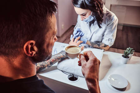A man receiving a manicure in the beauty salon, relaxing and drinking a coffee while a beautician master does manicure procedureの写真素材
