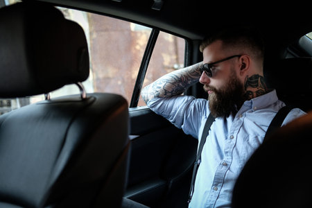 Serious bearded man is sitting in the car. He is wearing shirt, sunglasses and suspender. He has...