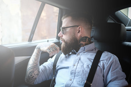 Imposing bearded man in sunglasses is sitting in the car. He is wearing shirt and suspender. He has...