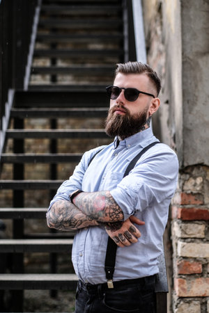Brutal bearded bully is standing near stairs at slam. He is wearing shirt, suspender and sunglasses....