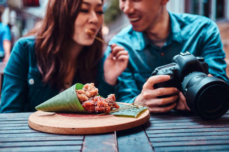 Happy young chinese couple sitting at cafe outside enjoying traditional asian food and watching pictures in photo camera.の写真素材