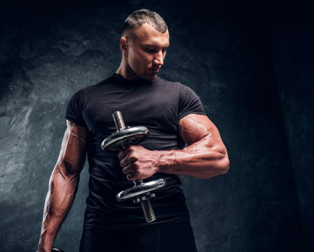 Muscular man is lifting dumbbells and looking at his muscules. There are dark background.の写真素材