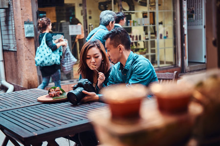 Happy young chinese couple sitting at cafe outside enjoying traditional asian food and watching...