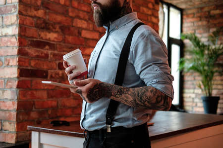 Bearded businessman is standing by the  table with tablet and coffee in his hands. He is wearing shirt and suspender. He has tattoes on his arms and neck. Office at background.の写真素材