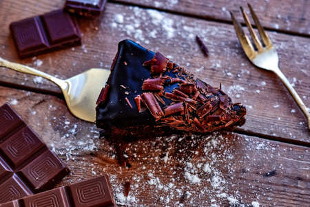 Delicious slice of chocolate cake with peaces of chocolate, fork, spoon and powdered sugar on dark table.の写真素材