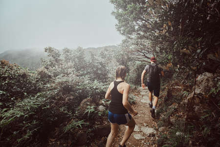 Young couple is running by the path in foresty mountains. There are green thickets and trees around. Fog and clouds at background.の写真素材