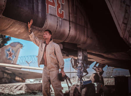 Young man is standing in a parking of military jets. He looks away. Man has a helmet in his...