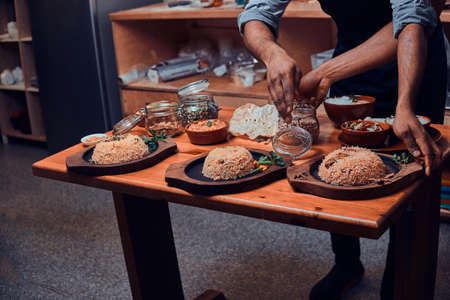 Cook picks up spices from jar for his indian dishes. There are few wooden plates with rice, jars with ingridients and papadoms on the table.の写真素材