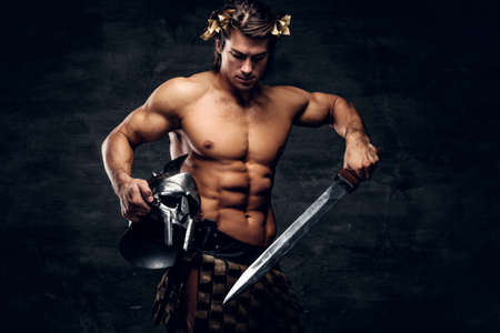Attractive powerful man in wreath is posing on grey background and holds helmet and sword in his hands.の写真素材