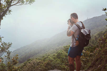 Young man with backpack is taking photo of beautiful nature on his photo camera while hiking in mountains.の写真素材