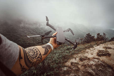 Man with tattoo is holding drone ready to depart. There are mountains in rainy fog at background.