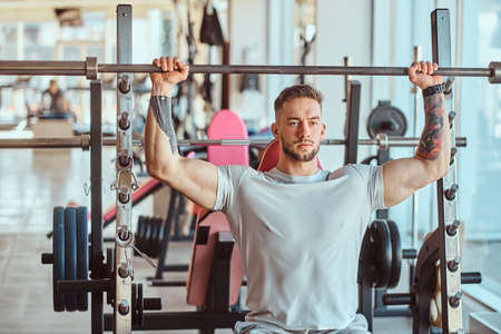Young tattooed bodybuilder is doing his workout with barbell in gym. There are many sport equipment around.の写真素材