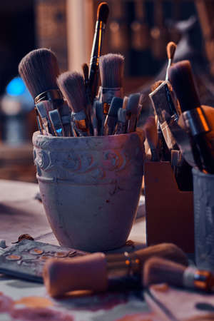 There are dark and a lot of different brushes on artists table in jars. There are artisticks mess on the table.の写真素材