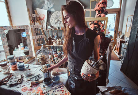 Talented artist in workshop is mixing paint for her new project while holding jar with brushes.