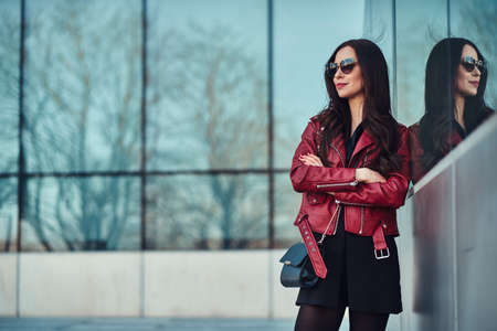 Attractive smiling woman in red jacket and sunglasses is posing near glass building.の写真素材