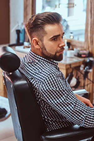 Attractive pensive man is waiting for his turn to get a haircut at busy barbershop.の写真素材