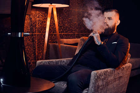 Pensive tattooed man is sitting on the armchair and starting smoke hookah. There are nice vapour.